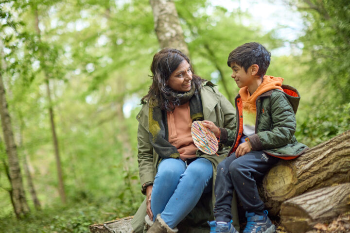 Woman and child holding a cardboard easter egg craft sit on a log in the woods